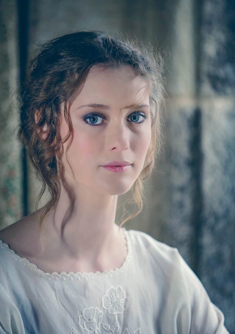 Elegant portrait of a young woman with blue eyes and curly hair, styled in a vintage-inspired white dress.
