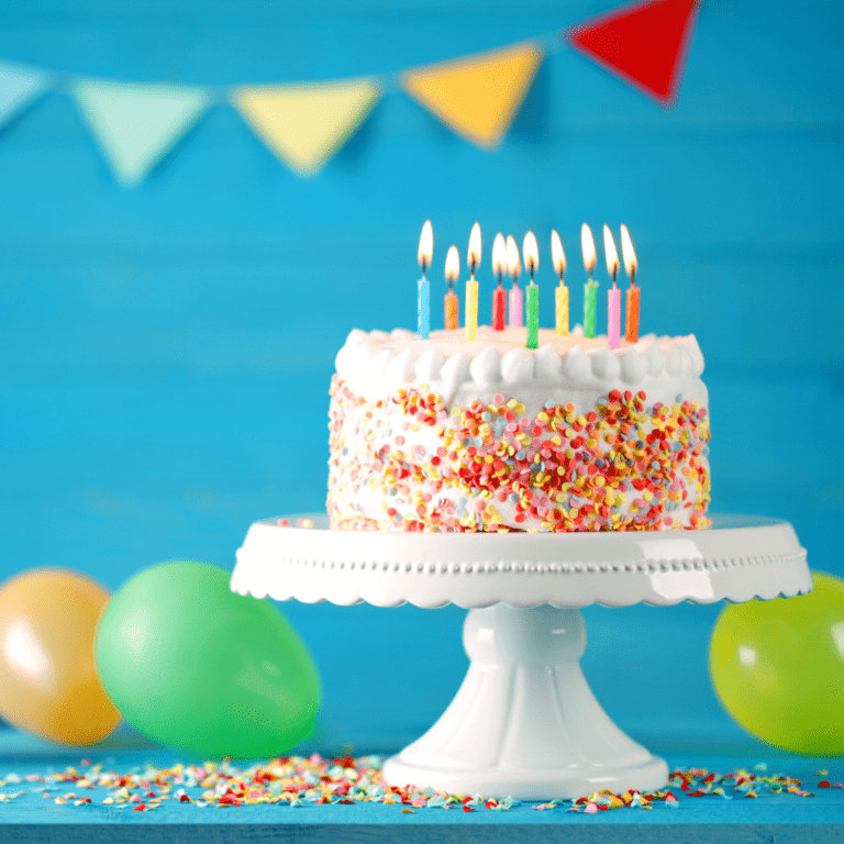 Birthday cake with colourful sprinkles and lit candles on a white cake stand, with balloons and bunting in the background.
