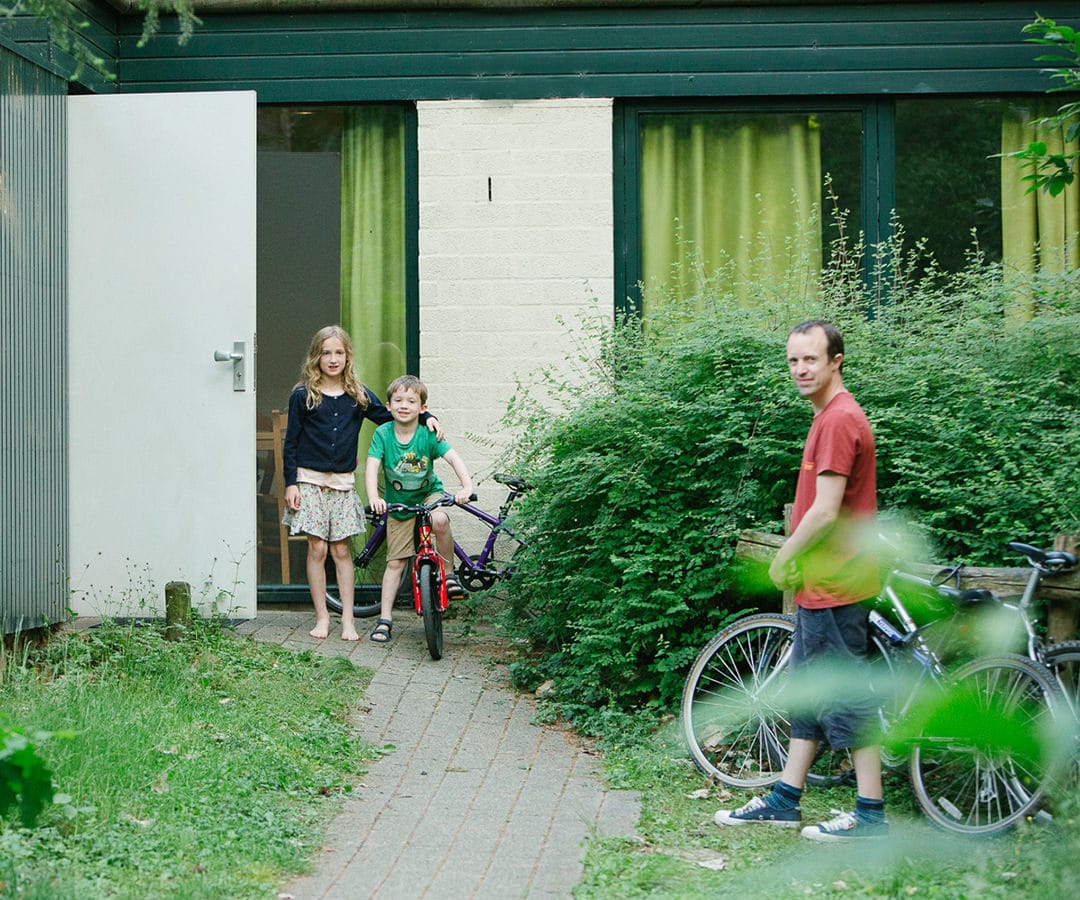 Children with bicycles outside a modern green and white house, family photography in a garden setting.