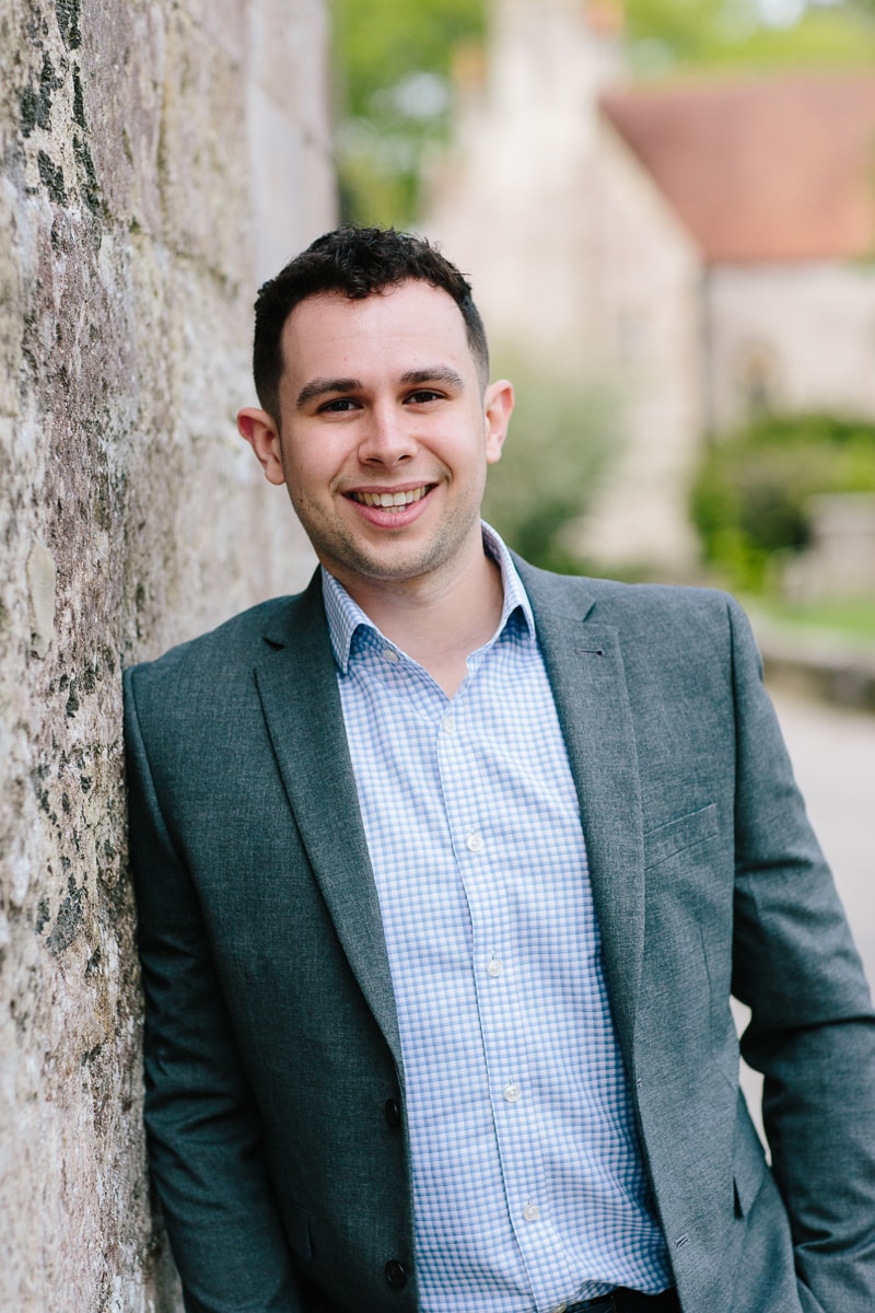 Young man in smart casual attire smiling against a rustic stone wall in an outdoor setting.