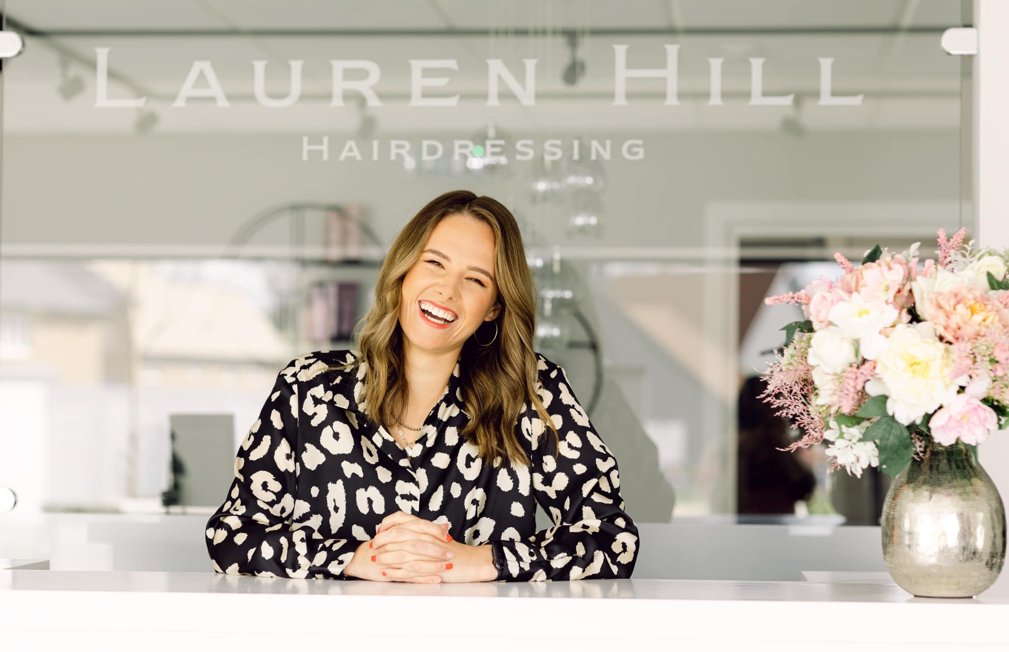 Stylish woman smiling at hairdressing salon reception, floral arrangement in foreground.