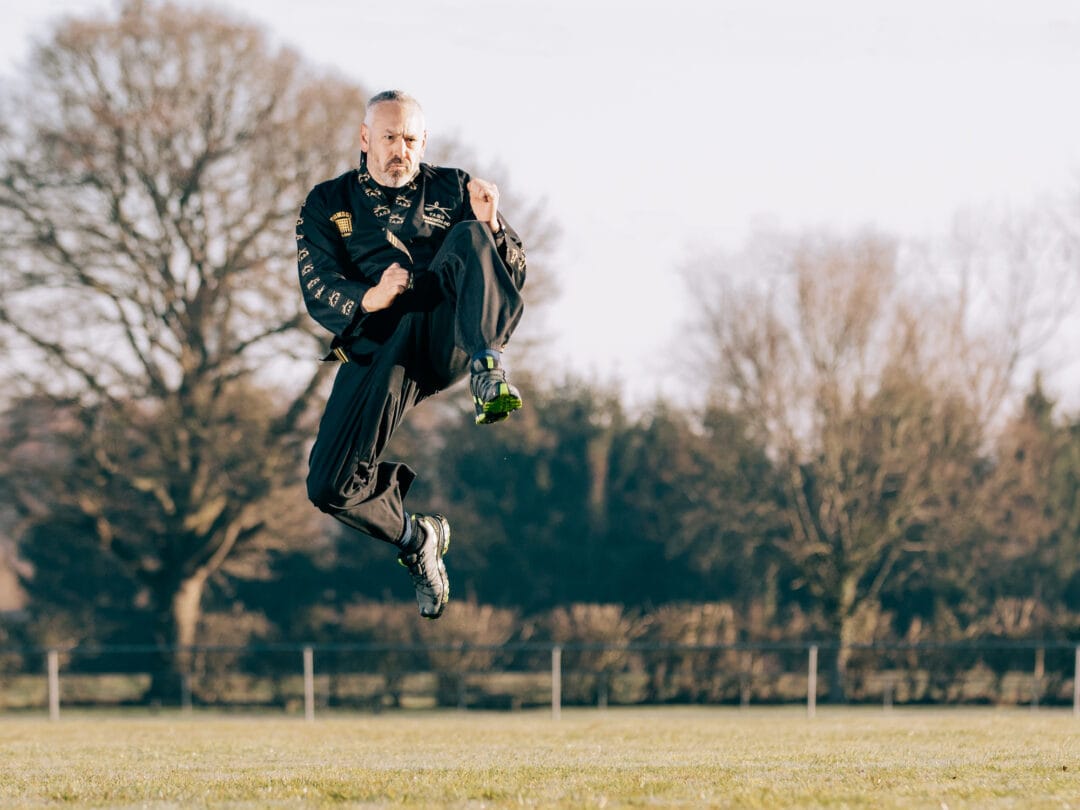 Dynamic sports photography of a man mid-air during a martial arts or athletic training session outdoors in natural light.