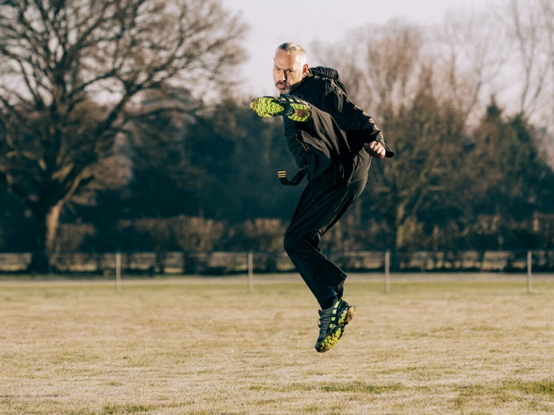 Dynamic action shot of a man jumping on a grassy field, wearing sportswear and football boots, captured by Jo Worthington.