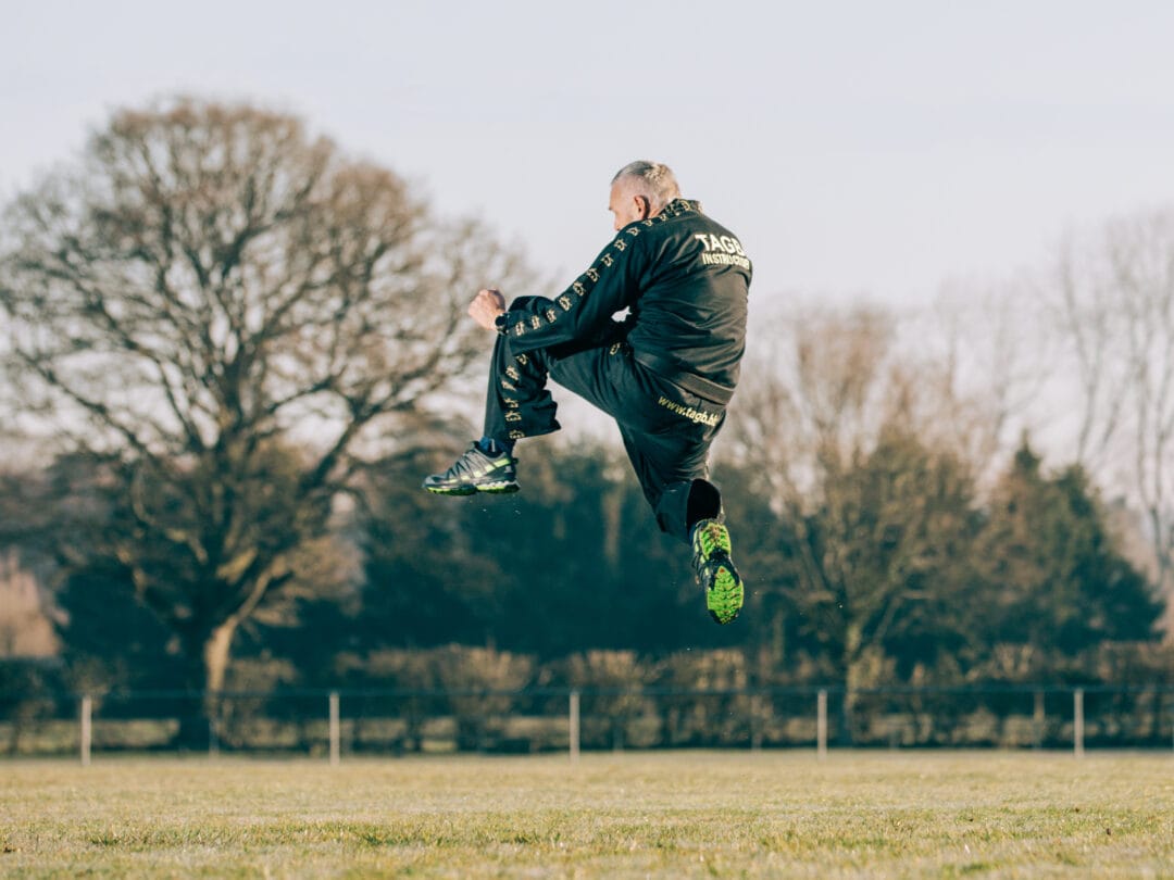Vigorous martial arts instructor performing a high kick outdoors in a park setting.
