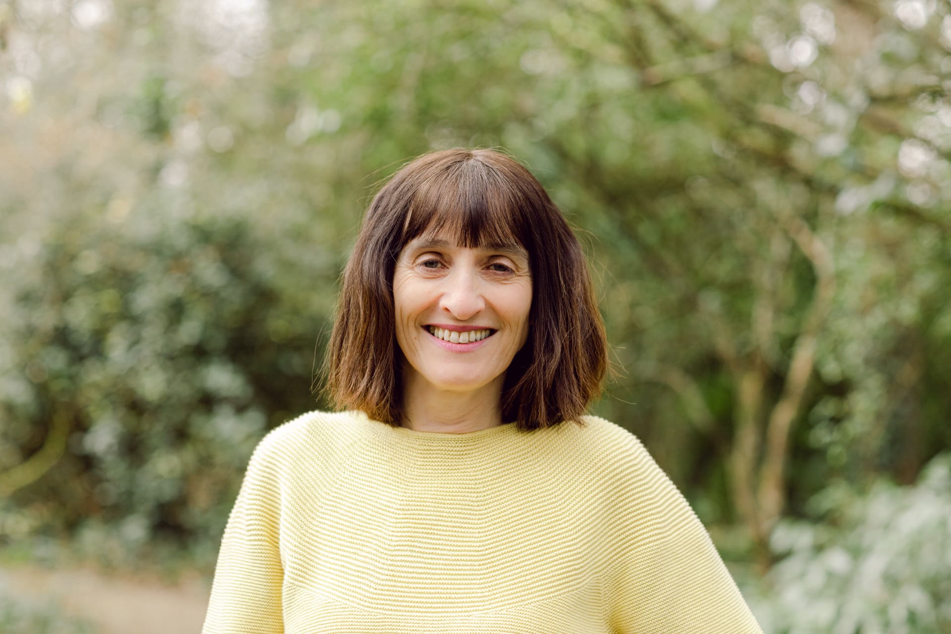 Natural outdoor portrait of a smiling woman with brown hair in a yellow top, lush green background.