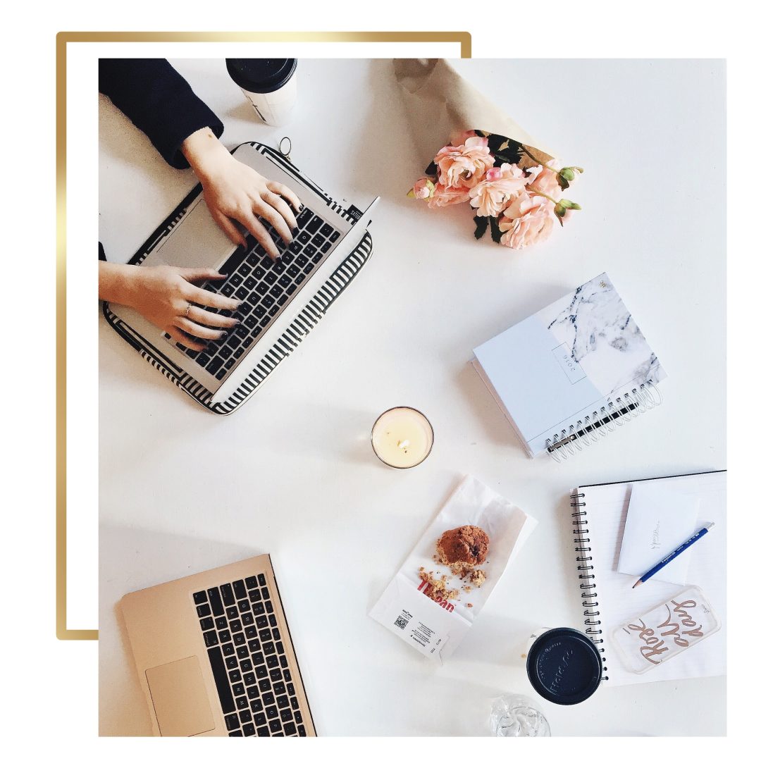 Bright workspace with laptops, notebooks, a candle, flowers, and snacks, perfect for creative work and photography.
