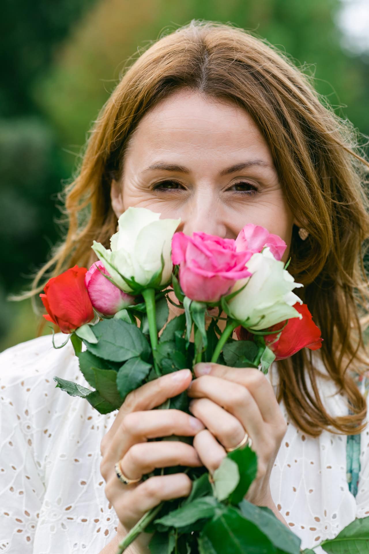 A woman with brown hair holding and smelling a colourful bouquet of roses outdoors in a garden.