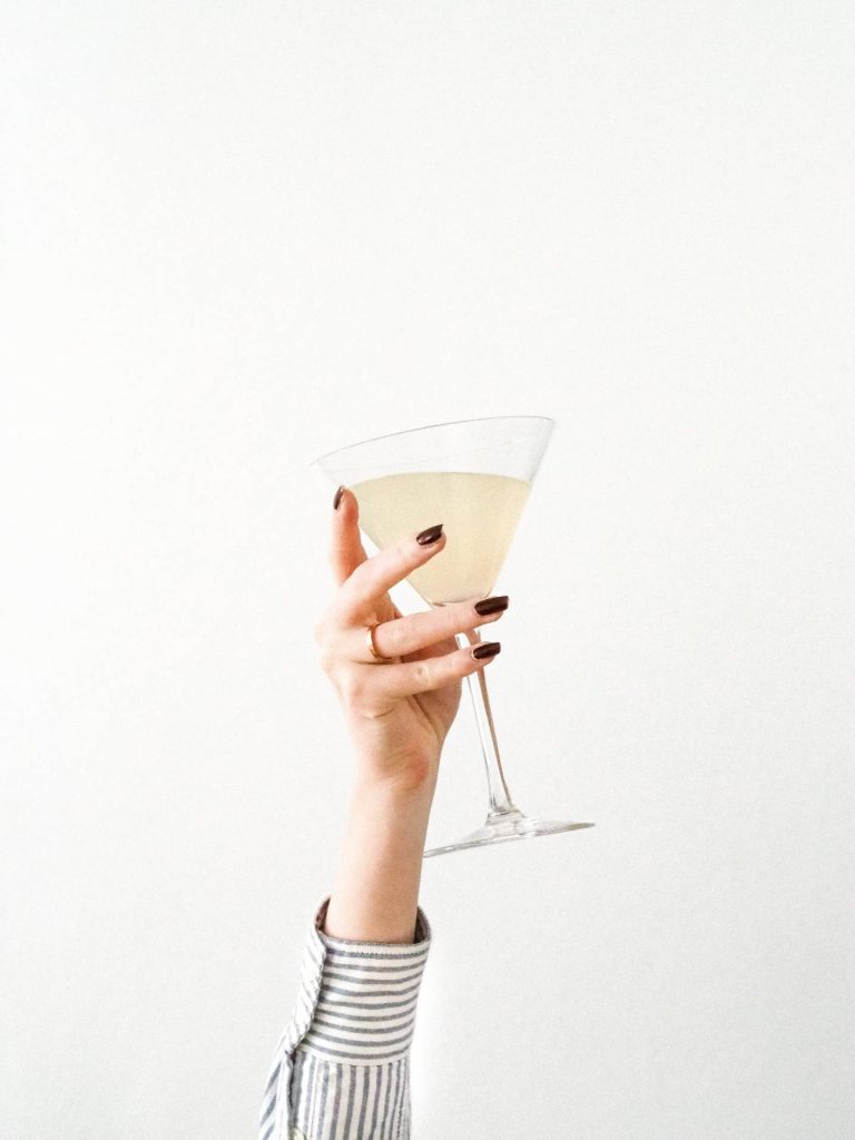 Elegant hand holding a cocktail glass with white wine or Champagne, minimalistic style, jewellery, striped shirt.